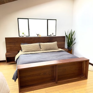 May include: A modern wooden bed frame with a dark walnut finish. The bed features a headboard with floating nightstands and a footboard with open shelving. The bedding includes a gray comforter and tan pillows.