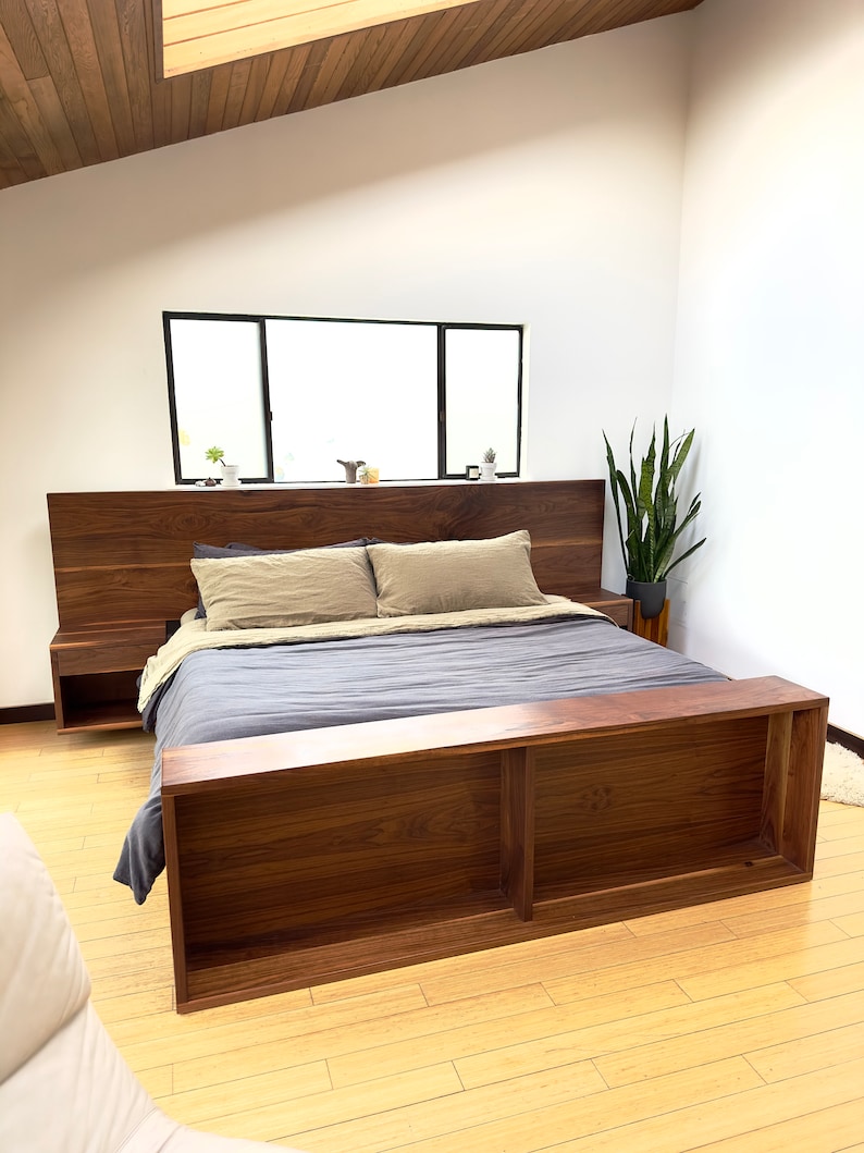 May include: A modern wooden bed frame with a dark walnut finish. The bed features a headboard with floating nightstands and a footboard with open shelving. The bedding includes a gray comforter and tan pillows.