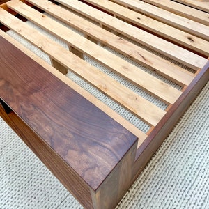 May include: A wooden bed frame with a dark brown headboard and a light brown slatted base. The frame is made of solid wood and has a modern design. The bed frame is shown on a white rug.