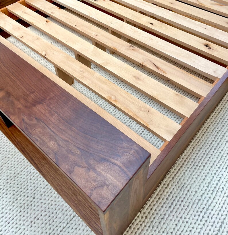 May include: A wooden bed frame with a dark brown headboard and a light brown slatted base. The frame is made of solid wood and has a modern design. The bed frame is shown on a white rug.