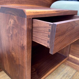 May include: A handcrafted wooden nightstand with a drawer partially open, showcasing dovetail joinery. The nightstand is made of dark brown wood with visible grain, and features a lower open shelf. The design is modern and minimalist.