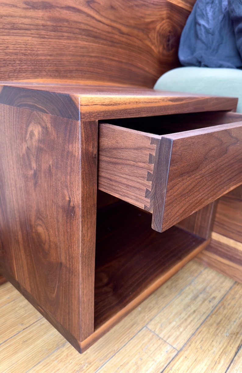 May include: A handcrafted wooden nightstand with a drawer partially open, showcasing dovetail joinery. The nightstand is made of dark brown wood with visible grain, and features a lower open shelf. The design is modern and minimalist.