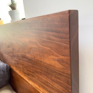May include: Close-up of a solid wood headboard with a rich, dark brown finish. The wood grain is prominent, showcasing natural patterns and textures. The headboard has a clean, modern design, perfect for a bedroom.