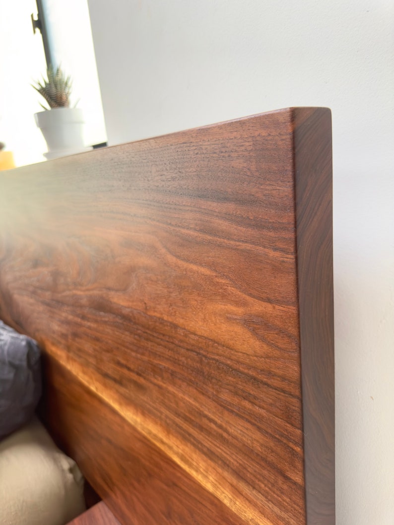 May include: Close-up of a solid wood headboard with a rich, dark brown finish. The wood grain is prominent, showcasing natural patterns and textures. The headboard has a clean, modern design, perfect for a bedroom.