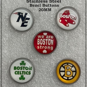 May include: A collection of custom-made, infused glass buttons with stainless steel bezels. The buttons feature various Boston sports team logos and text, including the Patriots, Red Sox, Celtics, and Bruins. Each button is 20MM in size.