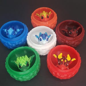 May include: A selection of brightly coloured 3D-printed dragon figurines in textured, round containers. The dragons and containers are in shades of blue, green, red, and orange. The background is a dark grey.