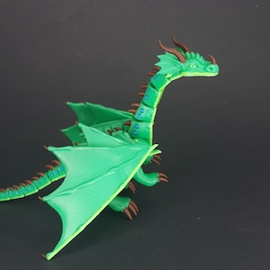 May include: A green dragon figurine with detailed wings, a long neck, and a tail. The dragon has brown horns and claws, with blue accents along its spine and tail. The figurine is set against a dark background.