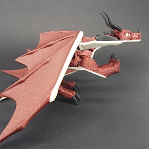 May include: A detailed figurine of a red dragon with large, textured wings. The dragon has a long tail, black claws, and horns. The wings are a darker shade of red with white accents. The dragon is posed on a dark surface.