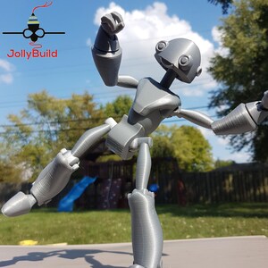 May include: A gray 3D printed robot figure with articulated limbs, posed in a dynamic action pose. The robot has a large head with two large eyes and a small mouth.