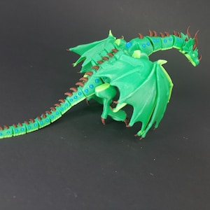 May include: A green dragon figurine with outstretched wings and a long tail. The dragon has brown accents on its spine, horns, and claws. Blue details are visible on the body. The figurine is set against a dark background.