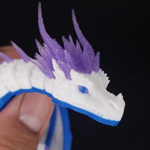May include: A 3D-printed dragon figurine with a white head, blue eyes, and purple spikes. The dragon's body is white and blue. The figurine is held in a person's hand against a dark background.