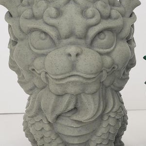 May include: A gray stone sculpture of a mythical creature with a dragon-like head and a body covered in scales. The sculpture has a detailed design and a textured surface.