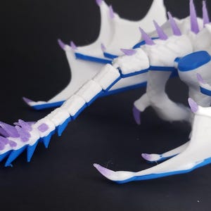 May include: A 3D-printed dragon figurine in white, blue, and purple. The dragon has a segmented tail with purple spikes and blue accents. Its wings and body are white with blue and purple details. The figurine is on a dark background.