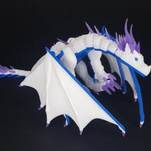 May include: A 3D-printed dragon figurine in white, blue, and purple. The dragon has large white wings with blue trim, a long tail, and sharp claws. The dragon is posed against a black background.