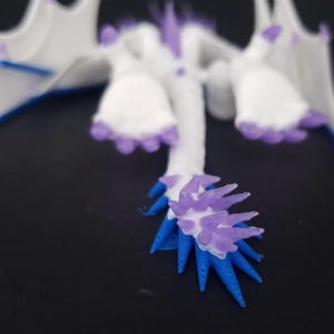 May include: A white and purple dragon figurine with blue accents. The dragon has outstretched wings, a long neck, and a spiky tail. The figurine is set against a dark background.