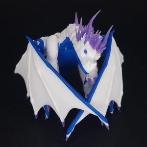 May include: A 3D-printed dragon figurine in white, blue, and purple. The dragon has large white wings with blue trim, a white body with blue accents, and purple spikes on its head. The dragon is posed with its wings spread.