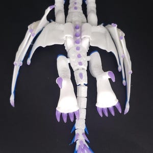 May include: A white and purple dragon figurine with articulated joints, wings, and a tail. The dragon has a detailed design with purple accents on the claws, spine, and tail. The wings have a delicate, webbed appearance. The figurine is set against a dark background.