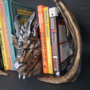 May include: A dragon head bookend in bronze, silver, and gold tones, holding several books. The bookend features detailed scales, horns, and a fierce expression. Books include titles like "The Steampunk User's Manual" and "Born a Crime".