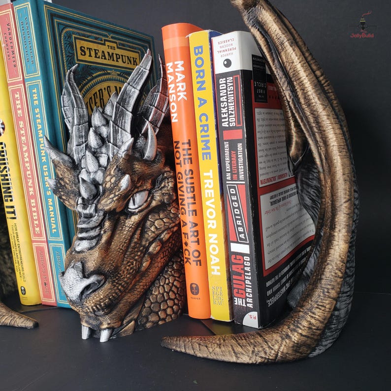 May include: A dragon head bookend in bronze, silver, and gold tones, holding several books. The bookend features detailed scales, horns, and a fierce expression. Books include titles like "The Steampunk User's Manual" and "Born a Crime".