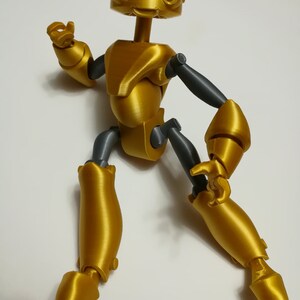 May include: A gold-colored articulated robot toy with gray joints. The robot has a large head with two large eyes and a small mouth. The robot is posed in a dynamic stance with one arm raised and the other arm bent at the elbow.