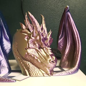 May include: A metallic dragon head sculpture with two large, purple and blue wings. The dragon head is gold and purple, with detailed scales and horns. The wings are curved and stand upright, creating a fantasy art piece.