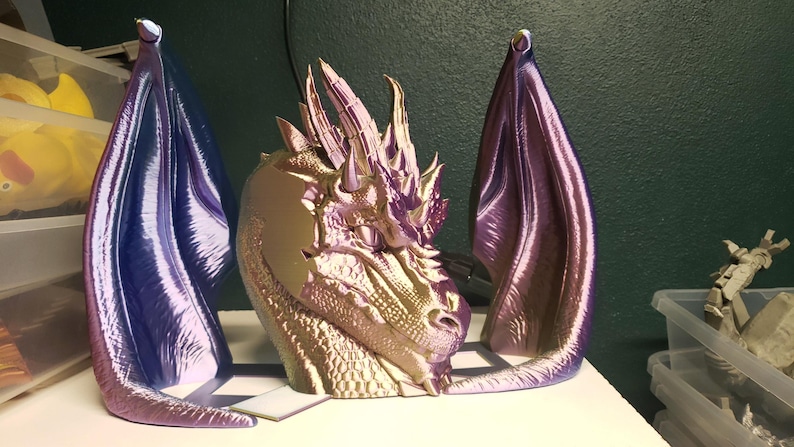 May include: A metallic dragon head sculpture with two large, purple and blue wings. The dragon head is gold and purple, with detailed scales and horns. The wings are curved and stand upright, creating a fantasy art piece.