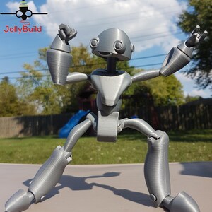 May include: A gray, articulated robot figure with a large head and large eyes. The robot is posed with its arms raised and its legs spread apart.