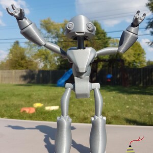 May include: A silver 3D printed robot with articulated limbs and a friendly face. The robot is posed with arms raised and a wide smile.
