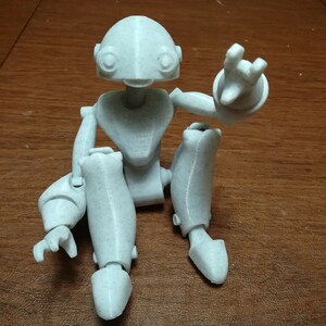 May include: A white, articulated, 3D printed robot figurine sitting with one arm raised. The robot has a rounded head with large eyes and a small mouth.