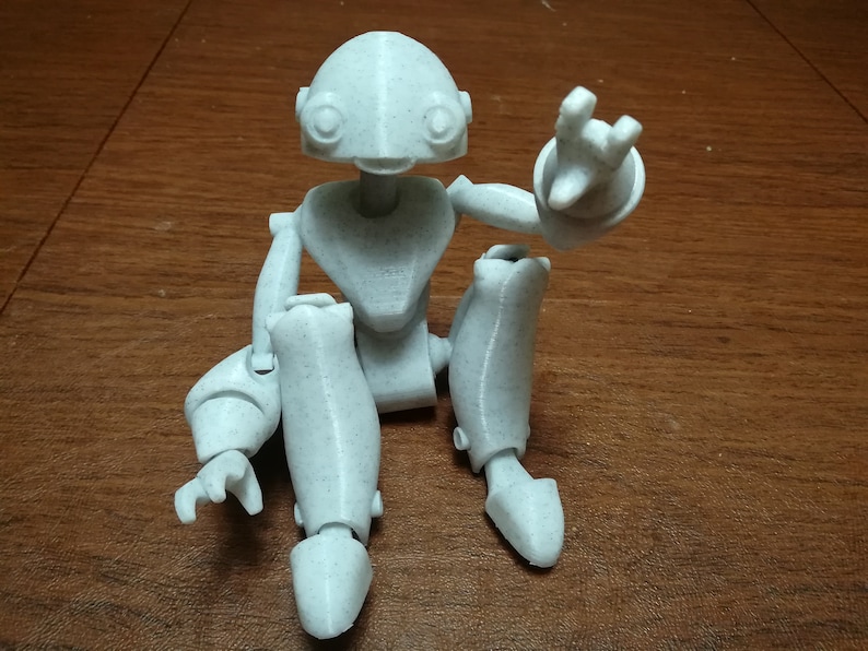 May include: A white, articulated, 3D printed robot figurine sitting with one arm raised. The robot has a rounded head with large eyes and a small mouth.