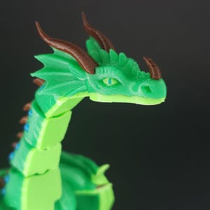 May include: A green articulated dragon figurine with brown horns and blue accents. The dragon's head is in profile, showcasing detailed scales and a focused gaze. The toy is made of plastic and is a fantasy-themed item.