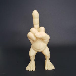 May include: A beige figurine of a hand giving the middle finger, with legs and feet. The figure has a rounded belly and is set against a dark background. The hand is the focal point, with the extended middle finger prominently displayed.