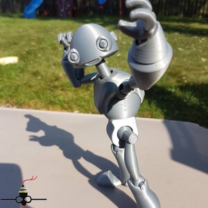 May include: A gray 3D printed articulated robot figure with a rounded head and large eyes. The robot is posed with one arm raised and the other arm bent at the elbow. The robot is standing on a gray surface.