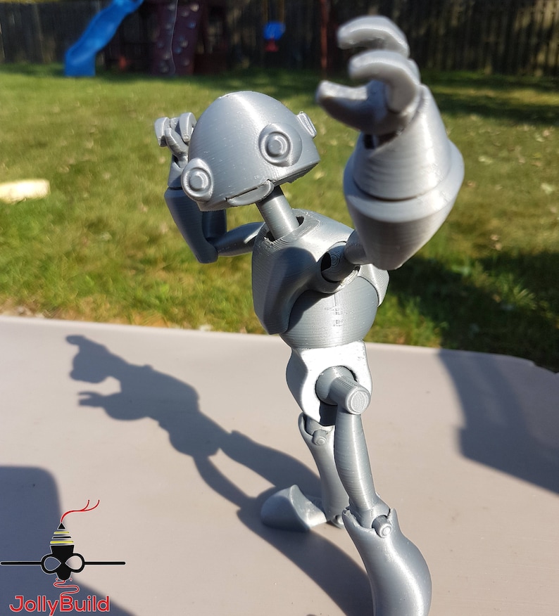 May include: A gray 3D printed articulated robot figure with a rounded head and large eyes. The robot is posed with one arm raised and the other arm bent at the elbow. The robot is standing on a gray surface.