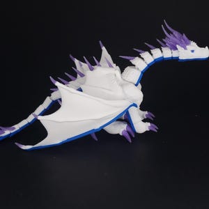 May include: A white dragon figurine with purple and blue accents. The dragon has large wings, a long tail, and detailed scales. The figurine is posed on a black surface.