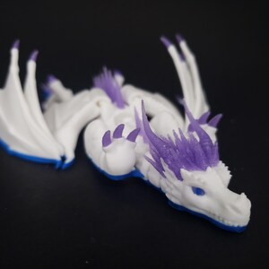 May include: A white and purple dragon figurine with blue accents. The dragon has detailed wings, horns, and claws. The figurine is lying down on a dark surface, showcasing its intricate design and color scheme.