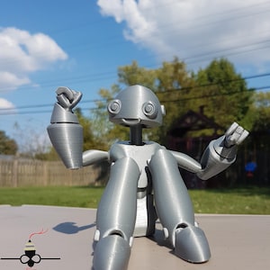 May include: A silver 3D printed robot figurine with articulated limbs, sitting with one arm raised and the other extended. The robot has a round head with large eyes and a small smile.