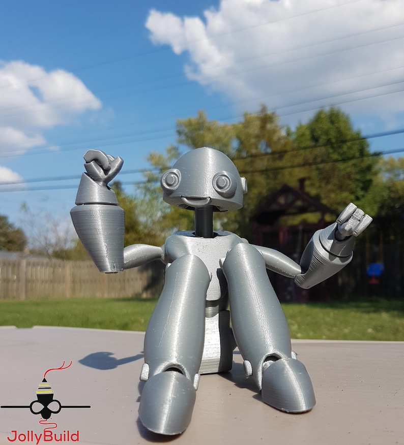 May include: A silver 3D printed robot figurine with articulated limbs, sitting with one arm raised and the other extended. The robot has a round head with large eyes and a small smile.