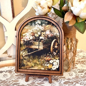 May include: A framed print of a wooden cart overflowing with colorful flowers. The print is set in a dark brown, arched wooden frame and displayed on a small wooden easel. The background features a vase of white flowers.