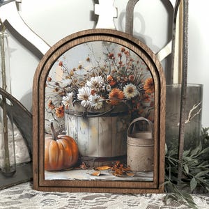 Può includere: Opera d'arte con cornice in legno a tema autunnale. L'opera raffigura una zucca, un secchio di fiori e un piccolo contenitore di metallo. La tavolozza dei colori comprende tonalità arancioni, bianche e marroni, evocando un'estetica autunnale rustica.