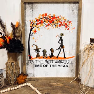 May include: A wooden sign with a white background and a black frame. The sign features a cartoon image of Jack Skellington, Sally, and Zero from the movie The Nightmare Before Christmas. The image shows them walking under a tree with orange and yellow leaves. The text on the sign reads "It's the most wonderful time of the year."