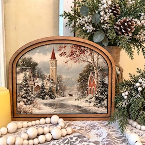 May include: A semi-circular wooden frame containing a winter scene print of a snow-covered village with a tall clock tower, red buildings, and evergreen trees. The print features a path, string lights, and a horse-drawn carriage in the distance.