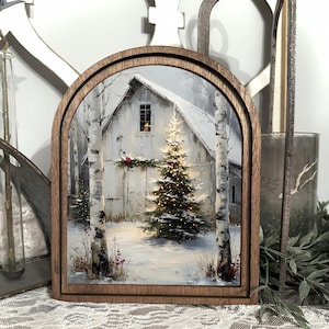 Könnte beinhalten: Gerahmtes Gemälde einer verschneiten Winterszene mit einer weißen Scheune, einem beleuchteten Weihnachtsbaum und Birken. Das Kunstwerk befindet sich in einem holzfarbenen, bogenförmigen Rahmen und schafft eine rustikale, saisonale Dekoration.