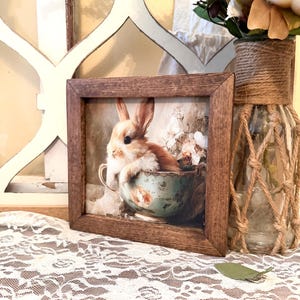 May include: A framed print features a brown and white rabbit nestled in a teal teacup with floral accents. The print is set in a rustic brown wooden frame. The image is displayed on a white lace tablecloth, with a vase of flowers in the background.