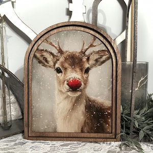 May include: A wooden framed artwork featuring a reindeer with a bright red nose. The reindeer is depicted in a snowy scene, with a soft, neutral background. The frame is a warm brown color, and the artwork has a vintage aesthetic.