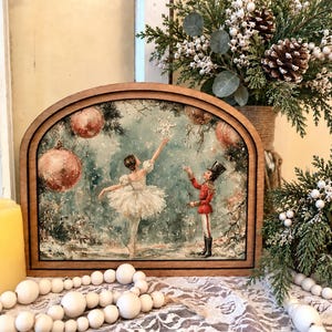 May include: An arched wooden frame containing a printed illustration of a ballerina in a white tutu and a nutcracker in a red uniform reaching toward a snowflake, surrounded by large red ornaments and a snowy, wintry forest background.