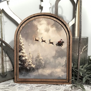 May include: A wooden framed Christmas scene showing Santa Claus in a sleigh pulled by reindeer. The artwork depicts a snowy forest and a cloudy sky. The frame has an arched top and a natural wood finish.