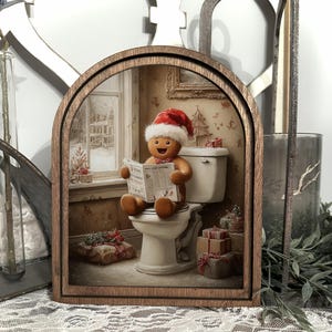 以下が含まれることがあります： 木製のフレームに入った、気まぐれなクリスマスをテーマにしたアート作品。サンタ帽をかぶったジンジャーブレッドマンが便器に座って新聞を読んでいる様子が描かれています。雪の降る窓、クリスマスプレゼント、お祝いの壁紙など、お祭り気分を盛り上げます。