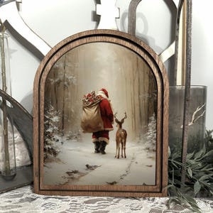 Puede incluir: Escena navideña enmarcada en madera que representa a Papá Noel con un traje rojo, caminando por un bosque nevado con un ciervo. La obra de arte está enmarcada en un marco de madera arqueado marrón oscuro.