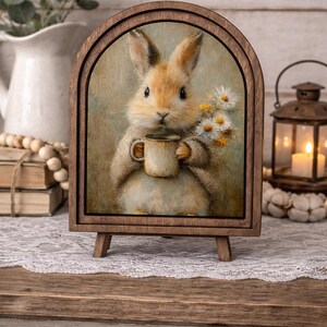 May include: A wooden framed print of a bunny holding a mug and flowers. The bunny is light brown with long ears and is holding a small brown mug. The print is displayed on a small wooden easel.